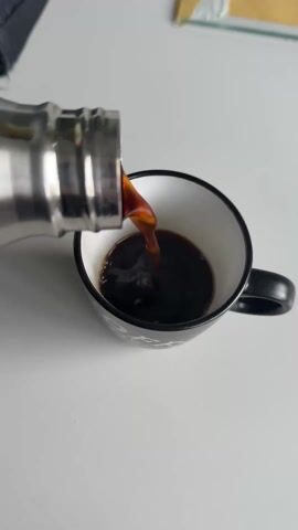 A close-up shot shows coffee being poured from a stainless steel thermos into a black and white mug. The coffee is dark brown and steaming slightly. The mug is on a white surface. A corner of a black bag is visible in the upper left, and a yellow envelope is partially visible in the upper right.