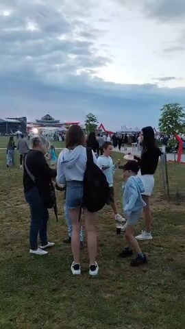 A group of people are gathered outdoors on a grassy area. In the foreground, several individuals, including women and children, are standing and talking. One woman in the foreground wears a light-colored hoodie and denim shorts, with a black backpack slung over her shoulder. A child in a light blue denim jacket and shorts stands near her. To the left, another woman in a dark jacket and jeans converses with someone partially obscured. In the background, a festival or event appears to be taking place, with tents, structures, and many more people visible. The sky is overcast with clouds. The overall atmosphere seems casual and social.