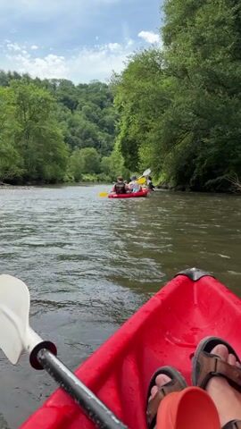 The video shows a view from a red kayak moving on a river. In the distance, several kayaks are visible, some grouped together near a rocky bank on the right side of the river. The river is bordered on both sides by dense green trees. The sky is partially cloudy. The videographer's feet, clad in sandals, are visible at the bottom of the frame, along with a kayak paddle. The water is somewhat choppy.