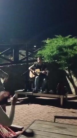 A man is playing an acoustic guitar on a small stage at night. He is wearing a dark t-shirt and jeans and is seated on a stool. There is a microphone in front of him. In the foreground, people are seated at tables, and some are clapping. In the background, there is a stone wall, a wooden fence, and trees. The scene appears to be an outdoor venue, possibly a restaurant or bar, with a casual atmosphere. The lighting is dim, and the overall mood is relaxed and festive.