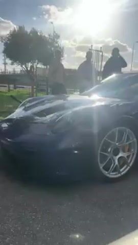 The video shows a collection of cars, including a blue sports car, a white Ford Mustang GT3 RS, and a red sports car. People are standing near the vehicles and walking in the background. The environment appears to be outdoors in a parking area with trees, a sidewalk, and street lights visible. The weather is sunny and partly cloudy. The video includes close-up views of the cars, highlighting their design and features, like the "GT3 RS" logo on the Mustang.