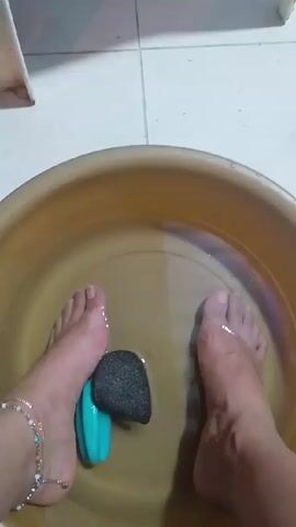 The video shows a pair of feet soaking in a tan-colored basin of water. In the water, there's a blue object and a dark gray, porous rock, possibly a pumice stone. The left ankle has a beaded anklet. The basin sits on a tiled floor, with a white plastic chair leg visible in the background. The camera angle is directly overhead. The background appears to be in a domestic setting.