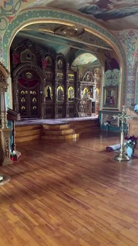The video shows the interior of a church. The foreground is a glossy wooden floor. Beyond that is a raised platform covered in a rug, and at the back, a large, ornate, wooden screen with depictions of religious figures. The screen has a set of double doors in the center. The walls and ceiling are decorated with frescoes and other religious artwork. The room appears to be empty and quiet.
