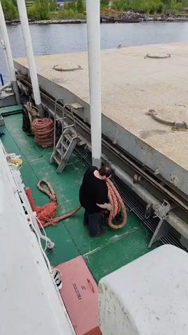 A person is seen on the green-painted deck of a boat, actively coiling a large, thick orange-brown rope onto a winding mechanism. The individual, wearing a black t-shirt and dark pants, is focused on handling the heavy rope. Several other coils of similar rope are visible on the deck near a metal ladder. To the right, a concrete pier runs parallel to the boat, with a body of water (likely a lake or river) extending into the background. Distant land with trees can be seen across the water. The scene appears to be under clear, bright daylight conditions. On the deck, in the foreground, there is a red rectangular box or hatch with the Cyrillic text "МЕШОК" (meaning "bag" or "sack") visible on its side. The boat's structure includes white vertical poles and railings. The setting is consistent with a dockside area in Medvezhyegorsk, Russia.
