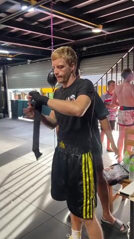 A blond male is wrapping his hands with black hand wraps in a gym setting. He is wearing a black t-shirt with a logo and black and yellow striped boxing shorts. A person with darker skin and hair stands behind him, looking on. In the background, other individuals are visible, some wearing athletic attire. The environment appears to be a boxing gym with gym equipment and garage doors. The lighting is artificial.