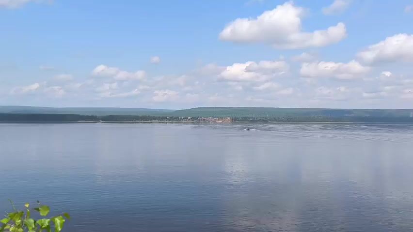 A wide, calm river under a bright blue sky with scattered white clouds. A distant shoreline is visible, covered in green trees, with a small cluster of buildings appearing as a village. A small boat is moving across the water, creating a wake. In the foreground, some green leaves are visible.