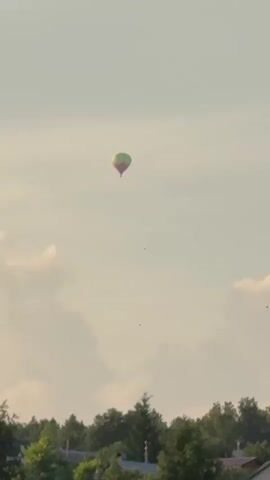 The video shows a hot air balloon in the sky over a rural landscape. The balloon is green and pink. The landscape includes trees, houses, and a body of water. The sky is cloudy and the sun appears to be setting. The video is taken from the ground.