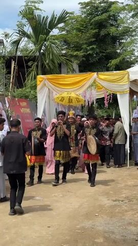 A group of young men in traditional black and gold attire are walking forward, playing musical instruments and a horn. One man in the center holds a yellow umbrella and wears a crown. They are walking under a yellow and white draped canopy. Other people are visible in the background, some watching the procession and others walking along with it. The setting appears to be an outdoor event, possibly a wedding celebration, with palm trees and a building visible.