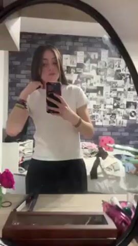 A young woman is recording herself in the mirror. She is wearing a white t-shirt and black pants. She has long, straight brown hair. She is touching her hair as she records. The bedroom is visible behind her, including a bed with clothes and a collage on the wall. There are pink flowers and a vanity in the foreground. The video seems to be taken with a phone.