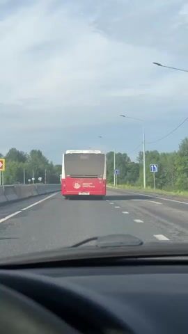 The video shows the view from inside a car driving on a road behind a red and white bus. The bus is the primary focus, with its rear facing the camera. The license plate "T205BC 159" is clearly visible on the bus. Above the license plate, there's text in Cyrillic which reads "ПЕРМСКИЙ ПЕРИОД НОВОЕ ВРЕМЯ" (Permian Period New Time), accompanied by a logo featuring a stylized animal head, possibly a mammoth or woolly rhino.

The road is asphalt with white lane markings. On the left side of the road, there are concrete barriers. Green trees and foliage line both sides of the road in the distance. Streetlights are present along the road, and a blue road sign indicating "ahead only or right turn" is visible on the right, along with a yellow triangular sign showing two-way traffic. The sky is overcast with clouds. The scene suggests daytime. Music is playing in the background throughout the video. The location is consistent with Perm Krai, Russia.