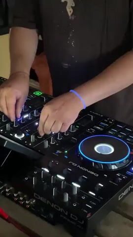 The video shows a person from the chest down, wearing a dark t-shirt with a graphic print, operating a DJ controller. They have a blue wristband on their left wrist. The DJ controller is black and features multiple knobs, faders, and illuminated platters. Red cables are connected to the controller. The person's hands are actively engaged with the controls, suggesting they are mixing music. The environment appears to be outdoors, possibly under a makeshift shelter, with wooden planks visible in the foreground and background. There are also some boxes and dark fabric structures in the background. The lighting is dim, with the illuminated parts of the DJ controller being the brightest elements. The video captures the DJ in action, focused on their equipment.
