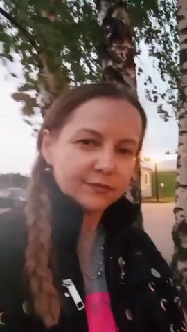 The video shows a woman with braided hair and a black jacket standing outside near some birch trees. She is looking at the camera and smiling. The background is blurry, but it appears to be an outdoor setting with a building and cars visible. The lighting suggests it may be dusk or dawn.