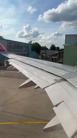 The video shows the wing of an airplane on a tarmac with buildings and trees in the background. The sky is blue with fluffy white clouds, suggesting it's daytime, likely late afternoon or early evening due to the warm lighting. The airplane's wing is white and prominent in the foreground, with visible flaps and structural elements. Part of the tail fin is visible on the left, displaying the pink "wizzair.com" logo. The ground is a concrete tarmac with a yellow line painted on it. The buildings in the background appear to be part of an airport facility, possibly hangars or terminal buildings, with one building painted in a light blue and another in green. The overall atmosphere is calm and suggests the moments before departure or after arrival. No people are directly visible in the frame. The time stamp indicates the video is very short, just a few seconds long.