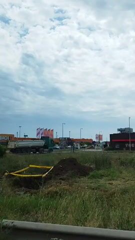 The video shows a construction site with yellow caution tape surrounding a dug-up area. A dump truck is parked nearby, and there are several buildings in the background, including what appears to be a fast-food restaurant with the words "YOUR WAY" visible on its side. Traffic signs are also present, indicating a speed limit of 30 and a roundabout. The sky is cloudy.