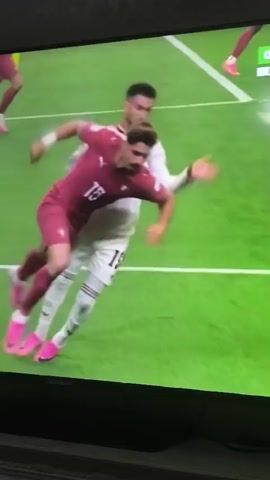 The video shows a football match on a television screen. Two male football players are visible on the green field. One player, wearing a maroon jersey and shorts, is on the ground in a defensive position, reaching out with his arm. Another player, wearing a white jersey and shorts, is in the process of kicking a white and green football towards the player on the ground. A third player, wearing a maroon jersey and shorts, is partially visible in the background on the right side of the screen. A graphic overlay at the top right corner of the screen reads "ФУТБОЛ 1" and "ПРЯМОЙ ЭФИР". The scene appears to be a live broadcast of a sporting event.