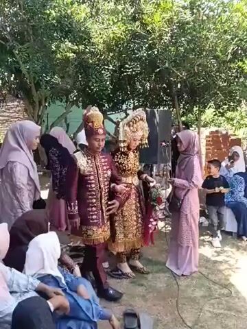 The video shows a wedding ceremony taking place outdoors. A couple, dressed in traditional wedding attire, are walking forward. The groom is wearing a maroon and gold outfit with a headdress, and the bride is in a gold and intricate dress with a large headdress. They are holding hands and walking past a group of people. Several women are present, wearing headscarves and modest clothing. Some are watching the couple, while others appear to be involved in the ceremony. There are trees and greenery in the background, and a white canopy is visible overhead. The ground appears to be dirt or gravel. There is a speaker on the left side of the frame. A small child is visible in the background, looking towards the camera.