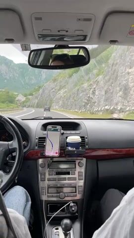 The video shows a view from inside a car driving on a highway. A smartphone, running a navigation app, is mounted on the dashboard. A paper cup of coffee sits next to it. The driver is visible only from the shoulders down, wearing a grey jacket and jeans. A passenger is on the right, visible only from the shoulder down, wearing a white shirt. The road runs through a mountainous area with light green vegetation. The dashboard is a light brown color with a wood trim. The car's stereo is gray.