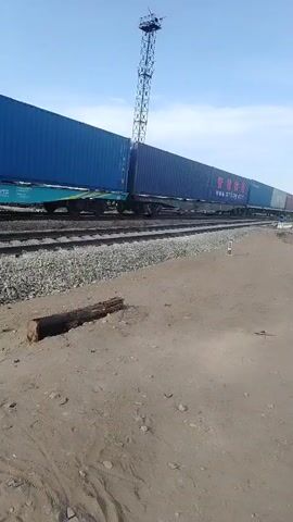 The media file shows a long train of blue shipping containers moving along a railway track. The train is traveling from right to left in the frame. In the foreground, there is a sandy and gravelly area with some debris, including a piece of wood. The railway track is clearly visible, with ballast supporting the rails.

In the background, a tall, lattice-like structure, possibly a radio tower or a signal mast, stands against a clear blue sky with some wispy clouds. The sun is shining brightly, suggesting it is daytime, likely late morning or early afternoon. The overall setting appears to be an open, possibly arid, area, consistent with the location context of Töretam, Kazakhstan, which is known for its steppe landscape and proximity to the Baikonur Cosmodrome.

The shipping containers are predominantly blue. Some of the containers have text visible. On several containers, the Chinese characters "智慧" (Zhìhuì, meaning wisdom or intelligence) are displayed prominently in red, along with the website address "www.smtlog.com" in white. Other containers show various identification numbers and markings, such as "94282498". There are no people visible in the media file. The activity depicted is the movement of the freight train, conveying goods across the landscape. The scene is industrial and functional, focused on transportation infrastructure.