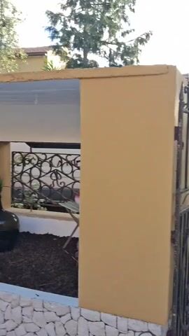 The camera slowly pans across the entrance to a courtyard. A wrought iron gate hangs ajar, revealing a gravel-covered drive leading to a yellow building with a red tile roof. Sunlight streams through the trees. The camera then moves to a small enclosed patio area, decorated with plants, a rustic wooden table, and chairs. A hanging lantern and a small cage complete the charming outdoor setting.