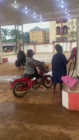 A person sits on a red motorcycle at a gas station. Another person stands to the right of the motorcycle, holding what appears to be a cup. Several other motorcycles and people are visible in the background. The ground is wet and appears to be dirt or mud, suggesting it may have recently rained. The gas station has a covered roof with lights, and buildings and trees are visible in the distance.