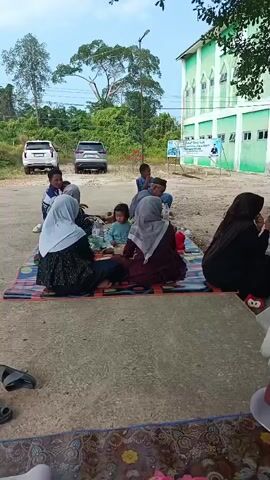 The video shows a group of people sitting on mats outdoors, possibly at a gathering or event. In the foreground, a colorful mat is visible, with a concrete area beyond it. A woman in a black hijab and black abaya is sitting on the right side of the frame, holding what appears to be a book or phone. In the mid-ground, several people, including women wearing hijabs and children, are seated on a patterned mat. They are facing away from the camera. To the left of the mid-ground group, two SUVs are parked. In the background, a green and white building, likely a mosque, stands with trees and vegetation surrounding it. A sign with text and an image of a person is posted near the building. The overall atmosphere appears to be relaxed and informal.