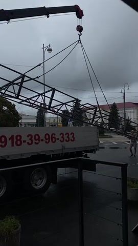 A crane is lifting a large metal truss structure off the back of a flatbed truck. The truck has the phone number 018-39-96-333 in red lettering on its side. Two men are standing near the truck, one watching the truss being lifted and the other appears to be walking away from the truck. The ground is wet, indicating it may have rained recently. The sky is overcast and cloudy. There are trees and buildings in the background, including what appears to be a street light.