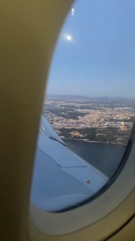 The video shows a view from an airplane window overlooking a city at dusk. The wing of the airplane is visible in the foreground, with the city lights twinkling in the distance. The sky is a deep blue, and the moon is visible as a bright disc. The city appears to be spread out along the coast, with a body of water separating it from the viewer. The lights of the city suggest a densely populated urban area. The overall impression is one of travel and the beauty of a city seen from above.