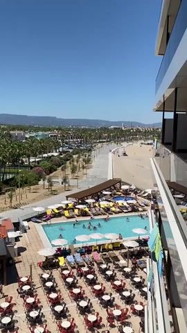 The video shows an aerial view of a resort or hotel complex on a sunny day. In the foreground, there is a large outdoor seating area with many red tables and chairs arranged in rows. Behind this area, there is a swimming pool with several people swimming and relaxing on lounge chairs under umbrellas. Beyond the pool, there is a sandy area that appears to be a beach, with palm trees and more lounge chairs. In the background, there are buildings and a distant view of hills and a clear blue sky. The overall scene depicts a lively resort atmosphere.