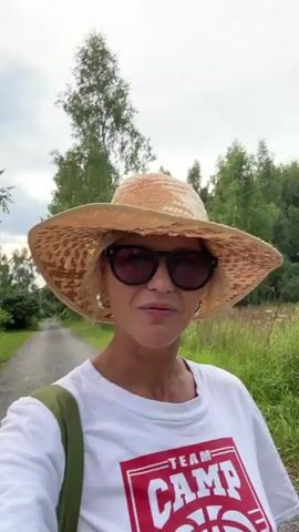 A group of people are walking down a gravel path lined with trees and greenery. The first person, wearing a straw hat and sunglasses, is talking to the camera. A second person, wearing a black t-shirt and a baseball cap, is walking next to them. Another person, wearing a blue shirt, is seen briefly in the background. The weather appears overcast.