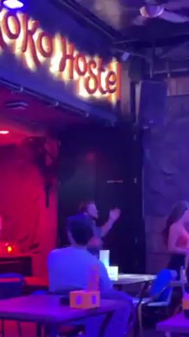 The scene is inside a bar called “Ko Hostel” with a neon sign. Several people are seated at tables, watching a man dance in front of a door labeled “STAFF ONLY”. A woman in an orange top and black skirt is also dancing. There’s a ceiling fan, speakers, and the lighting is red and purple.
