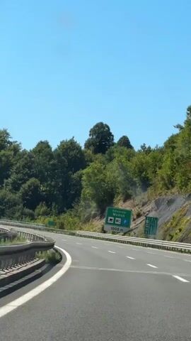 The video shows a view from inside a vehicle driving on a highway. The road curves to the left, with a guardrail on the left side. On the right side of the highway, there is a sign indicating "1000m" with icons for a restaurant, a restroom, and parking. Beyond the guardrail and sign, there is a steep, tree-covered hillside under a clear blue sky. Several vehicles, including a white truck and a white van, are visible on the highway in the distance.
