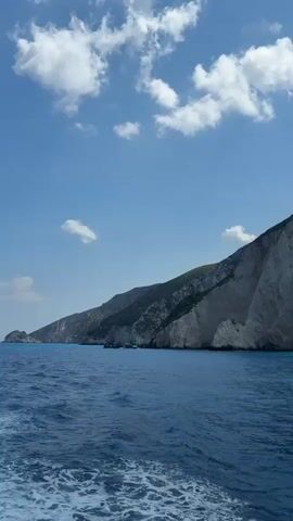 The video shows a boat moving on the ocean near a large cliff face. The water is a deep blue with whitecaps. The cliff is gray and rocky, with patches of green vegetation at the top. The sky is blue with scattered white clouds. A rope is visible in the foreground, suggesting the camera is mounted on a boat.