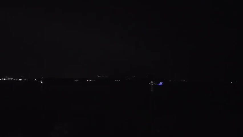 A wide shot shows a dark body of water at night. Lights are visible in the distance along the left side of the frame, suggesting a shoreline or a town. A single boat with blue lights is visible in the middle right of the frame, also on the water. The sky is completely dark, with no stars or moon visible. The scene is still and quiet, with no discernible movement other than the distant lights.