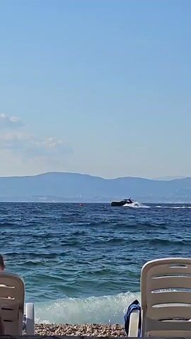 The video shows a view from a pebble beach looking out at the sea. Two white lounge chairs are visible in the foreground, one on the left and one on the right. A towel is draped over the right chair. The sea is blue and choppy with white waves breaking on the shore. In the distance, a dark-colored jet ski is moving across the water, leaving a white wake. Beyond the sea, a range of hazy mountains is visible against a clear blue sky with a few scattered clouds. The scene appears to be a sunny day at the beach.