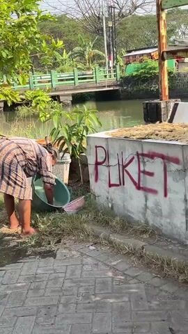 A person wearing a brown and white striped robe is bending down next to a body of water. They are holding a green bucket in their left hand and appear to be reaching into it with their right. In front of them, a white colander rests on the ground. To their right, a concrete structure is visible, with red graffiti on it. The person is barefoot. The environment appears to be outdoors, with vegetation and buildings in the background.