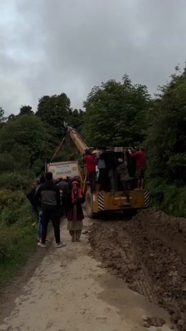 A group of people are gathered on a muddy dirt road, surrounded by lush green trees and foliage. A large yellow crane with a white box suspended from its boom is positioned on the road, with several individuals standing on the back of the crane and near the suspended box. The crane's boom is angled upwards, suggesting it is lifting or lowering the box. The road appears to be in a rural or mountainous area, with the sky overcast. The people are dressed in casual clothing. Some are looking at the box, while others are engaged in conversation. The scene suggests a logistical operation or a rescue effort.