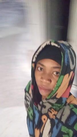 The video shows a person in a colorful hooded jacket and a black beanie, standing in a snowy environment. The person's face is visible, and they appear to be holding the camera and recording themselves. The camera moves around. At one point, the person is standing in front of a small, light-colored utility building. The overall scene is blurry due to the camera movement.