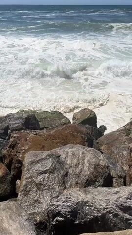 This media file captures a dynamic scene of ocean waves crashing against a rocky shore. The subjects are the natural elements: the vast ocean, the foamy white waves, and the large, weathered rocks in the foreground. The setting is a coastal area, specifically Torreira, Portugal, as indicated by the location context.

The activity depicted is the relentless action of the sea. Waves are actively breaking, creating white foam that surges and recedes around the rocks. The water appears to be a mix of deep blue and lighter green, with whitecaps indicating movement and power. The rocks are varied in color and texture, with some appearing darker and smoother, while others are lighter, rougher, and even tinged with green, suggesting the presence of algae or moss. The sunlight indicates it is daytime, likely mid-morning or afternoon, given the brightness. The weather appears to be clear and sunny, contributing to the bright, vibrant appearance of the water and the contrast with the shadows on the rocks.

Notable details include the scale of the rocks, which appear to form a breakwater or natural barrier against the sea. The foam of the waves creates a sense of energy and movement. The horizon line is visible, showing a clear distinction between the sea and the sky, which is a pale blue. There is no visible text within the media file itself. The overall impression is one of natural power and the raw beauty of the ocean at the coast.
