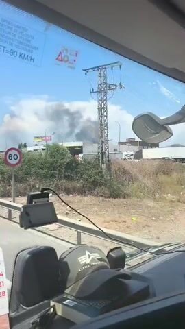 The video shows a view from inside a vehicle, looking out a windshield. In the distance, a large plume of black smoke is rising into a clear blue sky, indicating a fire. Below the smoke, a long, low-slung building is visible with signage. A red and white circular sign with the number 60 is on a pole in the foreground. The interior of the vehicle shows parts of a dashboard, including a black device, a cap with an alpha logo, and a calendar showing dates in July. The sky is bright and sunny, suggesting it is daytime.