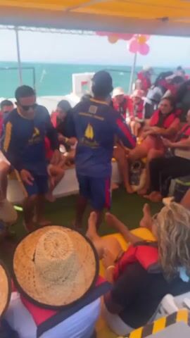 The video shows the inside of a catamaran filled with people enjoying themselves. A man in a navy blue shirt with “catamaran_mamalo” printed on the back is standing in the middle, appearing to lead a musical activity. Some people are seated on cushions along the sides, while others are more relaxed, lying down. Everyone is wearing life jackets. In the foreground, there's a straw hat. In the background, you can see the ocean and the sky. Pink and yellow balloons add to the festive atmosphere. The catamaran appears to be in motion.