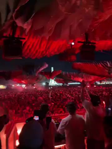 The video captures a dynamic night scene at a large outdoor music festival or concert. The perspective is from behind the DJ booth, looking out over a vast crowd filling an open field under a dark sky. The stage is intensely illuminated with red lights, which also highlight elaborate, layered structures suspended above. These structures, possibly abstract wings or organic forms, contribute to a dramatic and immersive stage design.

In the foreground, several DJs or performers are visible on the stage, primarily with their backs to the camera, actively engaging with their equipment. They are dressed in light-colored attire. The massive crowd extends far into the distance, appearing as a dense sea of people with many arms raised, indicating high energy and enjoyment. The overall atmosphere is vibrant and exciting, typical of a large-scale music event. The background suggests a clear night with no visible weather elements. The audio features energetic electronic dance music with a strong beat and vocals. No specific text or branding is discernible in the footage. The location context provided indicates this event is in Indio, United States.