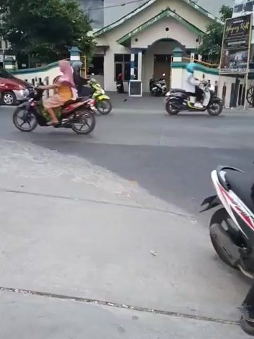 A street scene with multiple motorcycles and cars. In the foreground, a gray concrete surface is visible. Further back, several motorcycles are in motion. One motorcycle, ridden by a person in a white helmet and light-colored clothing, is traveling from left to right across the frame. Another motorcycle, ridden by someone in a dark helmet and dark clothing, is also moving from left to right. A third motorcycle, with two individuals on it, one wearing a helmet and the other without, is moving from left to right and partially obscured by the other vehicles. A white car is traveling from left to right in the mid-ground. Behind the motorcycles, a red car and a black SUV are parked or moving slowly. In the background, a residential building with a white facade and a green roof is visible, along with some trees and other structures. The overall scene appears to be a street in a populated area, likely during daylight hours, with moderate traffic.