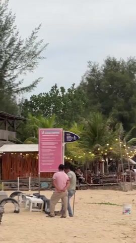 A vertical, full-shot, eye-level video captures a beach scene with a group of people and structures. The foreground is dominated by sand. Two men are in the center of the frame. One man, wearing a pink shirt and light-colored pants, stands with his back to the camera, facing left. The other man, in a green shirt and blue jeans, is to his right, also facing left, and is using a rake on the sand. To their left, near a white plastic chair, are several black tires stacked and leaning. A large pink sign with text in white is positioned behind the men, slightly to the left of center. To the right of the pink sign, a smaller blue sign with white text is visible. In the middle ground, several palm trees and other foliage create a backdrop. A low-slung structure with a thatched roof and bamboo-like walls is visible to the left, adorned with a string of lights. Further to the right, a similar structure with lights is also present, with several people seated at tables beneath it. The sky is overcast.