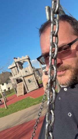 The video is a close-up shot of a man at a playground. He has short brown hair, a beard, and is wearing glasses and a dark blue shirt with a "CK" logo. He's holding onto the chain of a swing, looking directly at the camera. In the background, a child is swinging on another swing, and there is a wooden play structure resembling a castle. The playground has a red surface and is surrounded by grass and houses. The sky is clear and blue.