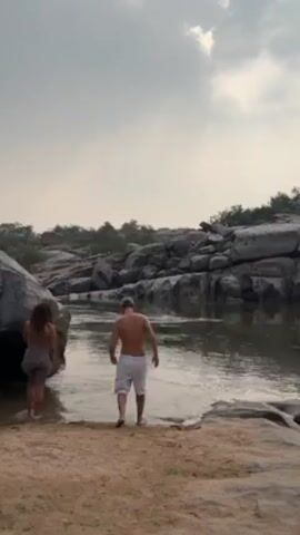 The video shows a couple wading into a river. The man is shirtless wearing white shorts and a baseball cap. The woman is wearing a dress or sarong. The river is surrounded by large boulders and greenery. The sky is overcast.
