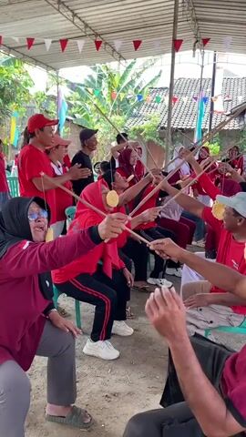 A group of adults, predominantly wearing red shirts or hijabs, are engaged in a game under a white canopy. They are seated on chairs and are holding wooden sticks vertically. At the end of each stick, a round, light-colored cracker is attached. The participants are attempting to eat the crackers by bringing them to their mouths. In the background, more people are observing the game, and colorful flags are strung across the canopy. The environment appears to be outdoors, with dirt ground and some foliage visible behind the canopy.