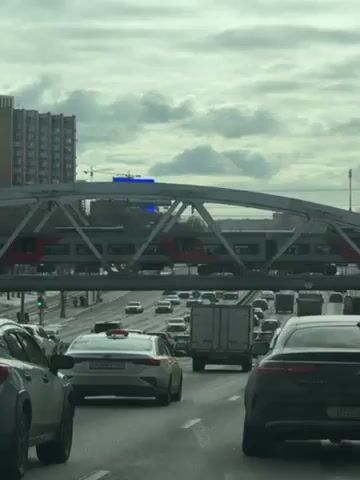 The video shows a view from inside a vehicle stopped in traffic on a multi-lane highway. Above the traffic, a light gray metal truss bridge carries a train with red and gray cars. The sky is overcast, and there's a large building visible to the left of the bridge. Cars of various makes and models surround the filming vehicle, with the closest cars being a white sedan, a white SUV, and a black sedan. Traffic is dense.