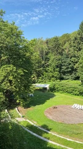 This media file shows an outdoor amphitheater setting, likely in Lübow, Germany. The scene is bathed in bright sunlight under a clear blue sky with a few wispy clouds, indicating it is daytime, probably late morning or early afternoon. Lush green trees form a dense backdrop, suggesting a natural, possibly wooded, environment.

The amphitheater itself features a circular wooden stage in the foreground, with a small pile of firewood in its center. Several white lounge chairs are arranged on the stage, suggesting a space for relaxation or perhaps a performance. To the left of the stage, a white canopy or tent is visible over a grassy area, with more white chairs scattered around it. The ground slopes down from the viewer's perspective, with tiered seating or pathways visible along the left side of the frame.

The overall impression is one of a serene, inviting outdoor space designed for gatherings or leisure. The greenery and sunlight evoke a feeling of peace and natural beauty. There is no visible text in the media file.