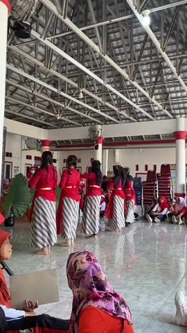 A group of six young women in traditional red tops and striped skirts are performing a synchronized dance in a brightly lit indoor space with a metal ceiling. They are facing away from the camera, and their movements are graceful and coordinated. In the foreground, two women are seated, facing the dancers. One woman in an orange hijab is looking at the dancers, and the other, also in a hijab, is looking down at a piece of paper. The background shows stacks of red chairs and a large green leaf prop. The floor is tiled and reflective, indicating a clean and well-maintained space. The overall atmosphere appears to be a cultural performance or event.