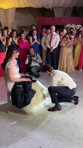 A man stands on his knees in front of a seated woman dressed in a wedding gown. The setting is festive, likely a wedding reception, with a crowd of guests in the background, many holding up their phones to record the moment. The man, wearing a white shirt, vest, dark trousers, and brown shoes, rises and gestures upward with a wad of what appears to be cash in his hand. The woman, seated on a wooden chair, smiles at him. The venue is decorated with fabric drapings and illuminated by soft lighting, casting a warm glow over the scene. A photographer kneels nearby, capturing the interaction.
